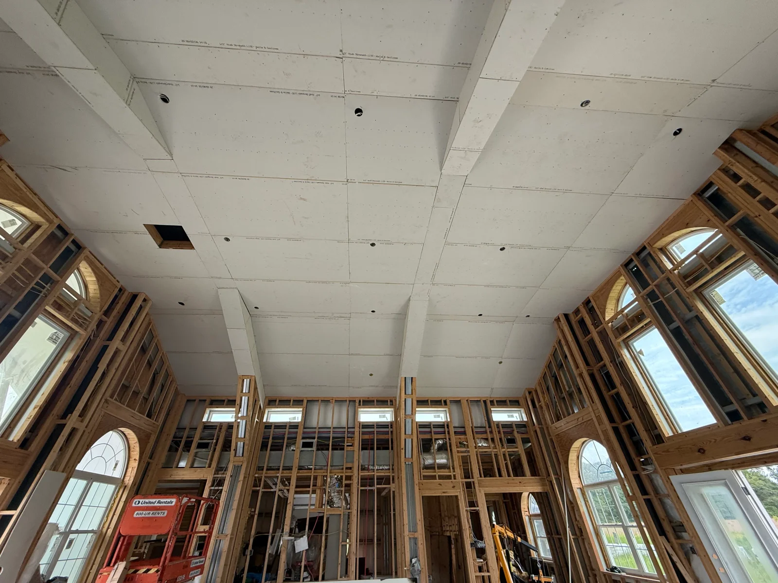 Drywall ceiling installation with recessed lights and wood window framing in open interior space